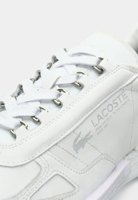Baskets en cuir blanc avec accents texturés, œillets argentés, lacets blancs, et un logo Lacoste subtil sur le côté.