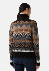 Vrouw met donker haar in een paardenstaart, gekleed in een hoog gesloten trui met zwart, bruin, grijs en witte zigzag- en geometrische patronen, gecombineerd met blauwe jeans.