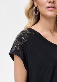 Czarna luźna bluzka z ozdobami z koralików i cekinów na ramieniu, wykonana z miękkiego, gładkiego materiału.