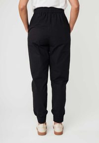 Schwarze Jogginghosen aus Baumwolle mit einem elastischen Bund und Bündchen; verfügt über eine glatte Textur und eine lockere Passform. Hellfarbige Schuhe sichtbar.