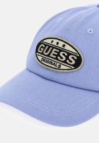 Lichtpaarse baseballpet met een ovale beige-zwart geborduurde "GUESS Originals" patch op het voorpaneel.