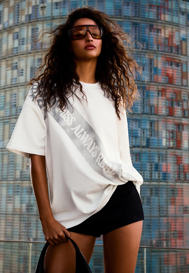 Femme aux cheveux bouclés portant un T-shirt blanc surdimensionné avec le texte « Class Always Real », short noir, grandes lunettes de soleil, tenant un sac noir, fond urbain.