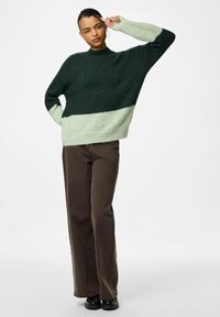 Maglione a blocchi di colore verde e verde chiaro con collo alto, vestibilità ampia e maniche lunghe, abbinato a pantaloni a gamba larga marroni.