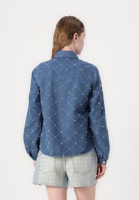 Blå denimskjorte med diamantmønster og hvite broderidetaljer, med lange ermer og krage, sett fra baksiden.