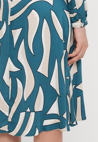 Tyrkysové šaty s púzdrovým designom, ktorý obsahuje abstraktné odtiene béžovej a bielej, čierne obrysy a jemnú, hladkú textúru. Viditeľné sú aj krátke rukávy s manžetami.