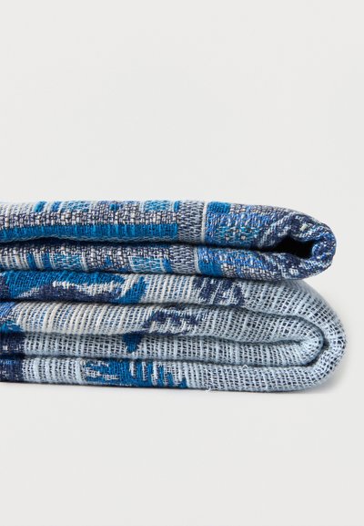 Deux couvertures tissées pliées avec des motifs géométriques bleus, bleu marine et blancs empilées sur une surface blanche.