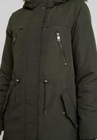 Olivgrön parkas med foder av päls i huvan, dragkedjor på bröstet och sidofickorna, dragsko i midjan och tryckknappar fram.