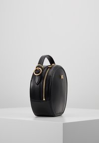 Kulatá černá kožená kabelka s horním uchem, zlatým zipem, kruhovým vzorem a zlatým logem na přední straně. Hladká textura.