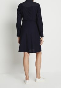 Abito a maniche lunghe di colore blu navy con vita aderente e gonna a campana. Tessuto strutturato, polsini con bottoni e dettaglio sul retro. Indossato con sneakers bianche.
