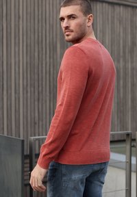 Homme en pull rouge et jean bleu regardant par-dessus son épaule, debout à l'extérieur près d'une rambarde en métal et d'un mur en panneaux de bois.