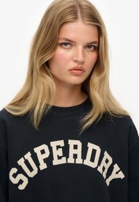 Sort sweatshirt med "SUPERDRY" trykt i cremefarvet bue-tekst. Blødt stof, rund halsudskæring, afslappet pasform og synlige sømadetaljer.