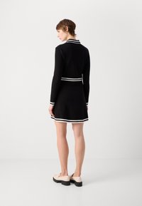 Robe noire à manches longues, col rayé blanc et taille cintrée. La jupe s'évase légèrement au-dessus des genoux, associée à des chaussures légères et un talon épais.