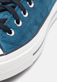 Κοντινό πλάνο μπλε σουέντ αθλητικού παπουτσιού με μαύρα κορδόνια, μεταλλικά πόρπες και λευκή λαστιχένια σόλα σε ανοιχτό φόντο.