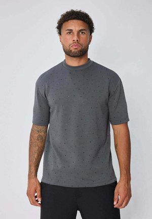 Homme aux cheveux bouclés et tatouages portant une chemise grise à manches courtes avec de petits points foncés et un pantalon noir, debout devant un fond uni.