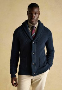 Marineblauer Strickcardigan mit Schalkragen, gerippter Textur, Knopfverschluss und zwei vorderen Taschen, getragen über einem karierten Hemd und einer Krawatte.