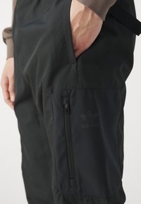 Svarta vadderade byxor med en sidodragkedjeficka och en diskret adidas-logotyp. Tillverkade av ett mjukt material, har de en avslappnad passform och en justerbar midja.