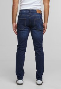 Hombre vestido con jeans ajustados de color azul oscuro y zapatillas blancas, de pie con los brazos relajados contra un fondo gris claro liso.