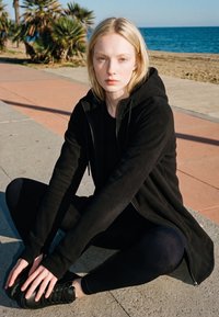 Mlada žena u crnoj sportskoj odjeći isteže se na otvorenom blizu plaže s palmama i oceanom u pozadini.