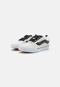 Adidași din suede alb cu șireturi negre, având o dungă laterală neagră, accente albe și o talpă din cauciuc texturată. Design de tip low-top.