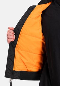 Ruka držící otevřenou černou prošívanou bundu, která odhaluje jasně oranžovou vycpanou podšívku proti černému oblečení.