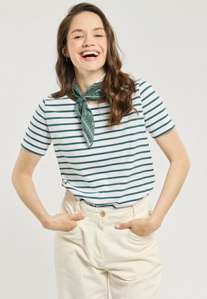 Femme souriante les mains dans les poches, portant un pantalon blanc, une chemise blanche à rayures vertes, et un foulard vert autour du cou sur un fond uni.