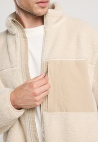 Beige fleece jacket with a zip front, soft texture, and a contrasting tan pocket. Features a high collar and lined interior.