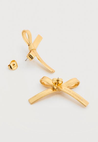 Σκουλαρίκια καρφάκια σε σχήμα φιόγκου από μεταλλικό υλικό με χρυσό τόνο και πίσω μέρος πεταλούδα, το ένα σκουλαρίκι δείχνει την μπροστινή όψη, το άλλο την πίσω με χαραγμένη την επωνυμία.