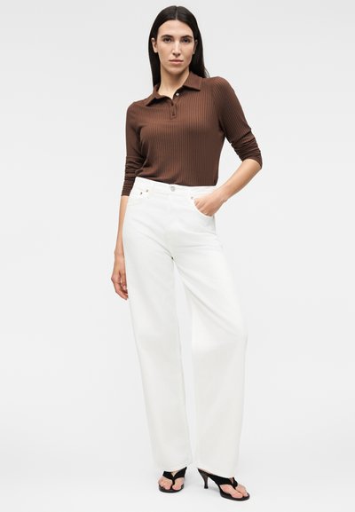 Γυναίκα με μακριά σκούρα μαλλιά φοράει καφέ ριμπ μπλούζα πόλο με μακριά μανίκια, λευκό παντελόνι με φαρδιά γραμμή και ψηλή μέση, και μαύρα πέδιλα με τακούνι, στέκεται με το χέρι στην τσέπη.