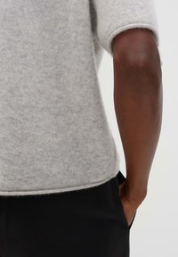 Bras d'une personne à la peau foncée portant un pull léger gris clair à manches courtes et un pantalon noir, la main reposant dans une poche.