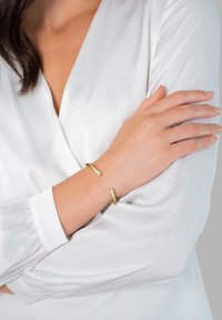 Bracelet jonc en or avec une finition lisse et polie, mettant en valeur deux accents en cristal clair. Porté sur un bras contre une blouse blanche.