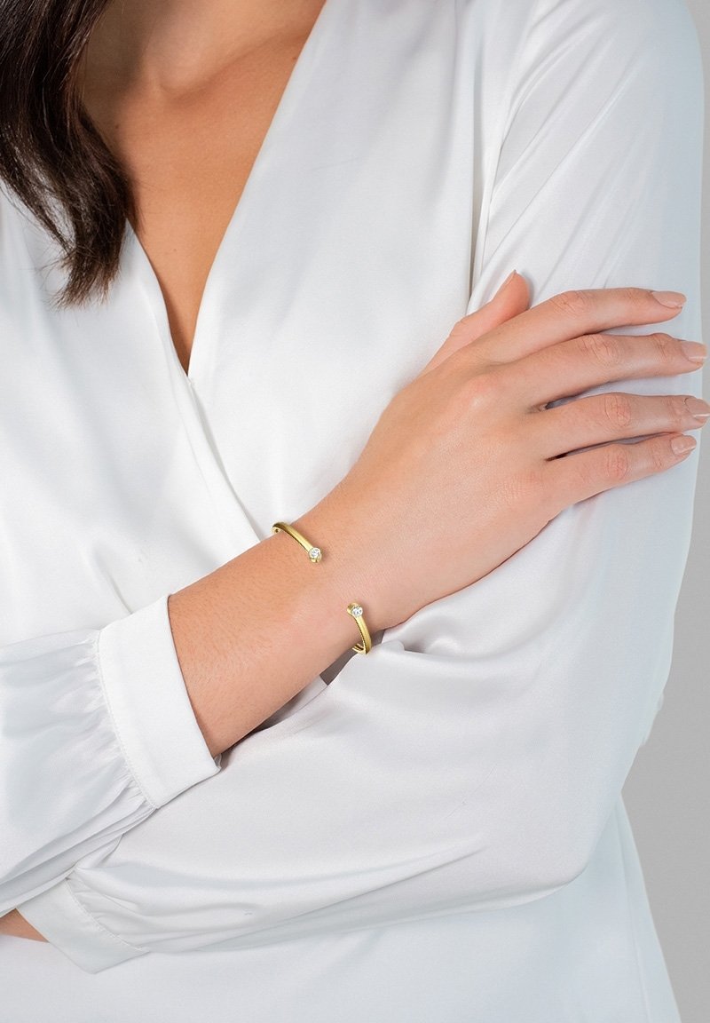 Bracelet jonc en or avec une finition lisse et polie, mettant en valeur deux accents en cristal clair. Porté sur un bras contre une blouse blanche.
