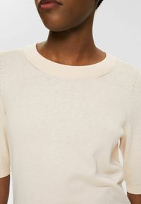 Top lavorato a maglia beige chiaro con maniche corte e scollo arrotondato, caratterizzato da una trama liscia e dettagli di cucitura discreti.