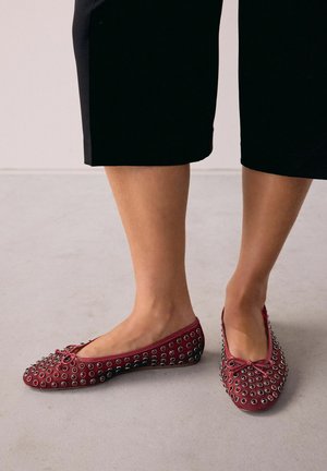 Gambe che indossano pantaloni neri a vita alta e ballerine rosse decorate con numerosi occhielli argentati, in piedi su un pavimento grigio chiaro.