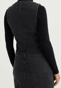 Person som bär en åtsittande svart denimväst över en svart långärmad topp och en matchande svart denimkjol med synliga bakfickor.