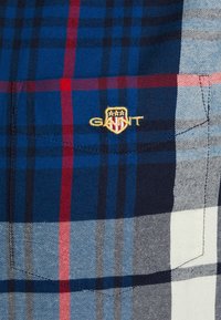 Camisa de cuadros con tonos azules, marinos y blancos, y acentos rojos. Incluye el logotipo bordado de GANT en el lado izquierdo. Tela texturizada.