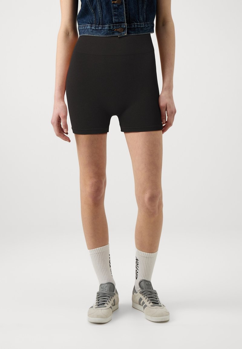 Femme portant un short côtelé noir taille haute, des chaussettes blanches avec texte noir, des baskets grises et blanches, et un gilet en jean court.