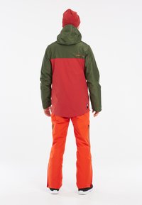 Herenwaterdichte jack in rood en groen, gecombineerd met oranje broek. Heeft een capuchon en ritszakken. Zachte textuur, sportieve pasvorm.