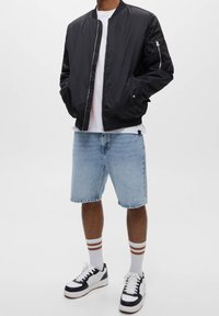 Veste bomber noire avec une fermeture éclair à l'avant, associée à un short en denim bleu clair, un t-shirt blanc et des baskets noires et blanches.