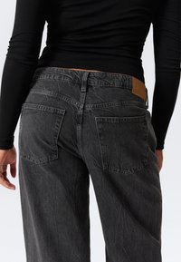Jeans negri din denim cu un finisaj texturat, având două buzunare pe spate și o etichetă din piele maro la talie.