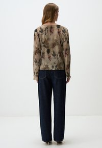 Langärmlige Bluse mit floralem Design in gedeckten Farben, aus strukturiertem Stoff gefertigt. Getragen mit hochtaillierten dunklen Jeans.