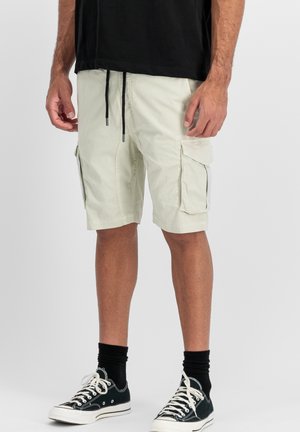 Light beige cargo shorts with two side pockets, drawstring waist, and a casual fit. Worn with black socks and black sneakers.