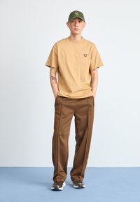 Tan cotton t-shirt with a small heart emblem, paired with brown sweatpants. The outfit is complemented by a green cap and sneakers.