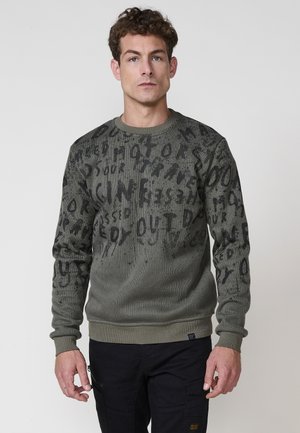 Maglione verde a maglia con tessuto testurizzato, caratterizzato da bold scritte nere. Polsini e orlo a costo, scollatura arrotondata. Indossato sopra pantaloni scuri.