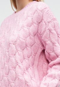 Jersey rosa de punto con cable que presenta un patrón texturizado con elementos redondeados, cuello redondo y mangas ligeramente holgadas. Material suave y peludo.