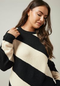 Pull à rayures noir et crème avec un col rond, texture douce, manches longues et poignets côtelés. Le tissu semble épais et chaud.