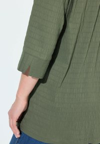 Olivgrüne Bluse aus strukturiertem Stoff, Dreiviertelärmel mit einer Schlitze und geradem Saum. Getragen mit Jeans.