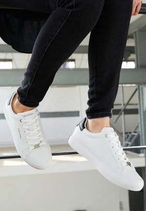 Osoba nosząca białe trampki Calvin Klein z czarnym detalem na pięcie i czarne spodnie, siedzi ze stopami uniesionymi w pomieszczeniu.