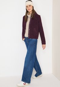 Bordowa kurtka wyściełana polarem z zatrzaskami, zestawiona z jasnobeżowym golfem i szerokimi niebieskimi dżinsami. Białe trampki z czerwonymi akcentami.