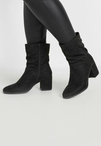 Botas de tobillo de ante negro con un diseño arrugado, cierre de cremallera y un tacón ancho. Punta afilada y textura suave.