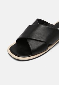 Sandale en cuir noir avec des sangles croisées, semelle plate et bord clair. Texture lisse avec des bords arrondis, conçue pour être enfilée.